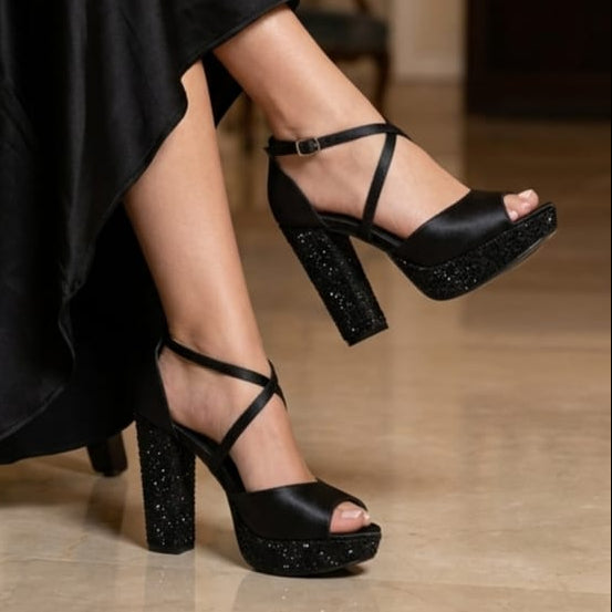 A pair of black platform high-heel sandals with ankle straps and glittery block heels, shown on a model wearing a black satin dress in an indoor setting.