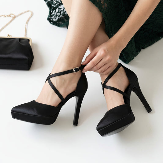 A person wearing black platform high-heeled pumps with ankle straps, adjusting the strap on one shoe. A black handbag with a gold chain is partially visible on the left, and a dark green lace dress is shown on the model against a clean white background.