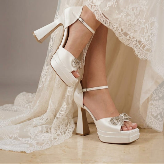 A pair of bridal high-heeled sandals worn with a white lace wedding dress. The shoes are off-white satin with platform soles, block heels, ankle straps, and jeweled embellishments on the toes.