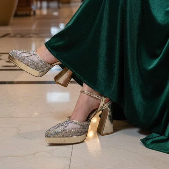 Person wearing a green dress and gold shoes in an indoor setting with a chandelier and plants.