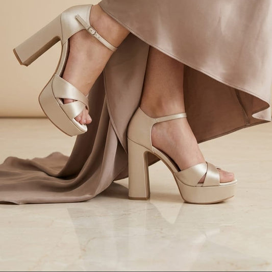 A pair of women's platform high-heel sandals in a light champagne satin, shown on a model wearing a flowing taupe satin dress. The sandals feature criss-cross toe straps, a single ankle strap with a small buckle, and a chunky block heel.