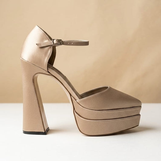 A single platform high-heel shoe in a light champagne beige satin-like material, featuring a block heel, open toe, and ankle strap with a small buckle, set against a warm beige background.
