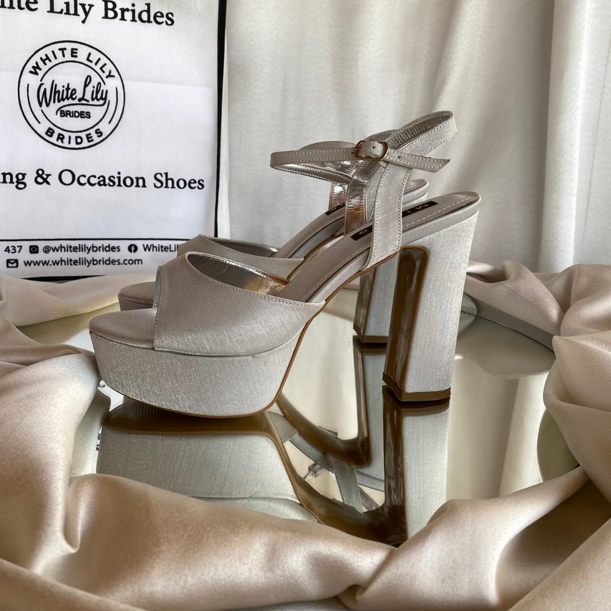A pair of White Lily Brides platform high-heeled sandals in a silver satin finish, displayed on a reflective surface with draped champagne silk fabric and a branded shopping bag in the background.