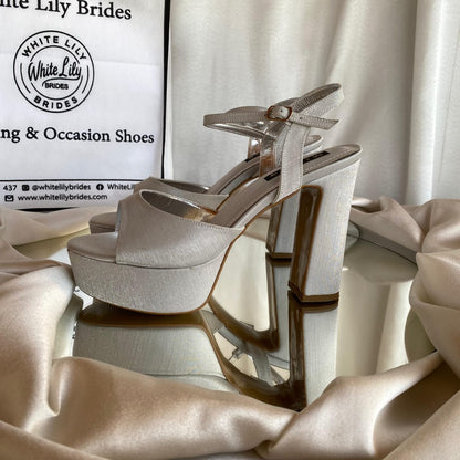 A pair of White Lily Brides platform high-heeled sandals in a silver satin finish, displayed on a reflective surface with draped champagne silk fabric and a branded shopping bag in the background.