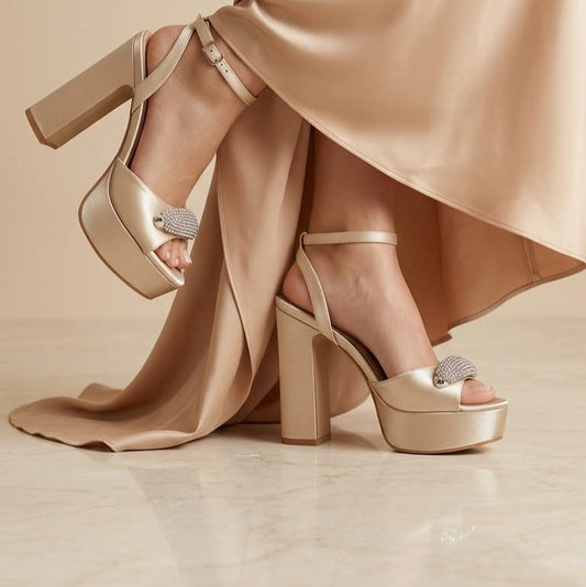 A pair of silver champagne gold platform sandals with block heels and rhinestone-embellished toe caps, shown on a model wearing a matching satin dress against a beige background.