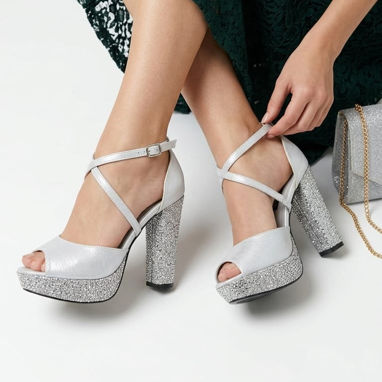 A woman wearing a dark green lace dress sits on a white floor, adjusting a pair of silver platform sandals with glittery silver heels and straps. Beirut