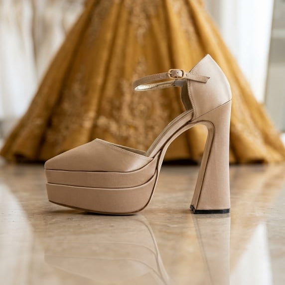 A Nude Champange beige platform high-heel shoe with an ankle strap, displayed in the foreground on a polished floor with a blurred gold gown on a mannequin in the background.