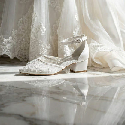 White lace bridal shoe with a low block heel and ankle strap featuring a pointed toe, posed next to a wedding veil.
