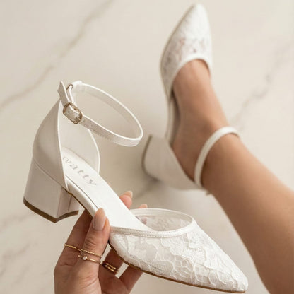 White lace bridal shoe with a low block heel and ankle strap featuring a pointed toe.
