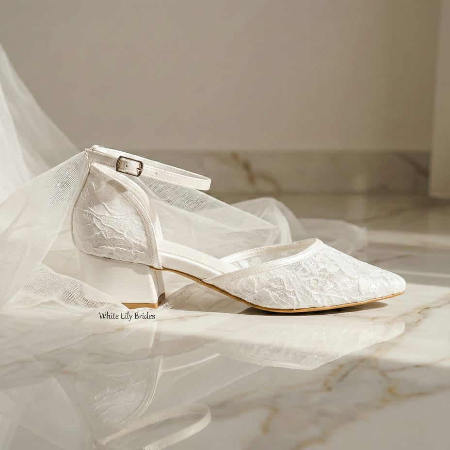 Side view of White Lily Brides ivory lace wedding shoes with a low block heel and ankle strap, placed on a marble floor with tulle fabric.