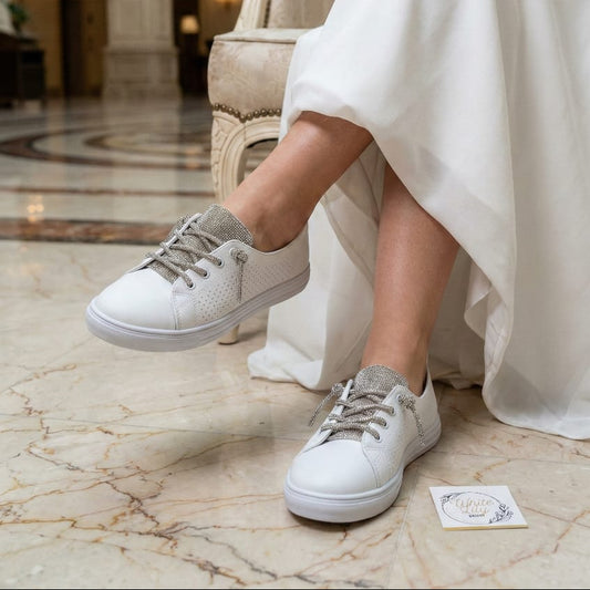 white bridal sneakers featuring sparkly crystal-encrusted laces, worn by a bride in a flowing wedding dress sitting on a chair in a luxury marble lobby.