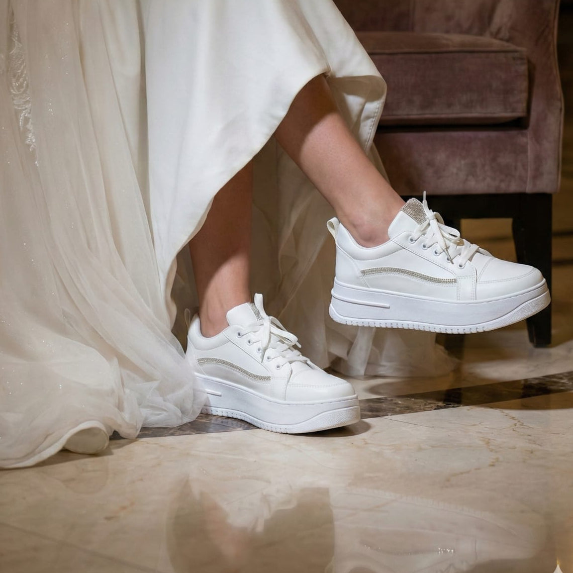 Side view of white platform bridal sneakers with crystal trim, worn by a bride in a luxury wedding dress sitting on a marble floor in a hotel lobby.